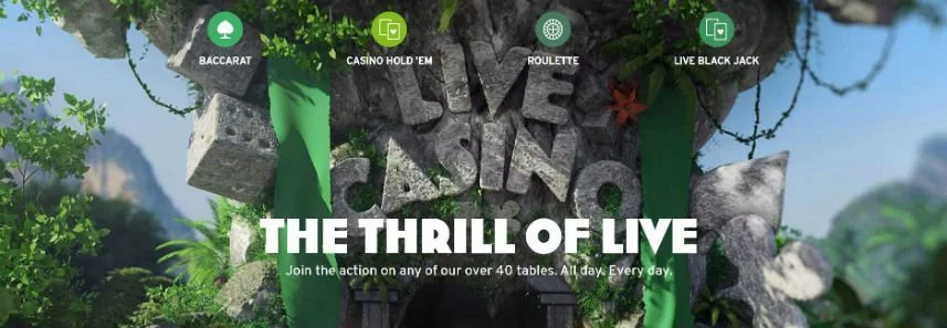 Bilden visar en djungel där ett stort träd(?) har stenar formade som tärningar hängandes och även stenar som formar ordet Live Casino. Under live casino står det "The thrill of live. Join the action on any of our over 30 tables. All day. Every day." Över ser vi länkar till Baccarat, Casino hold